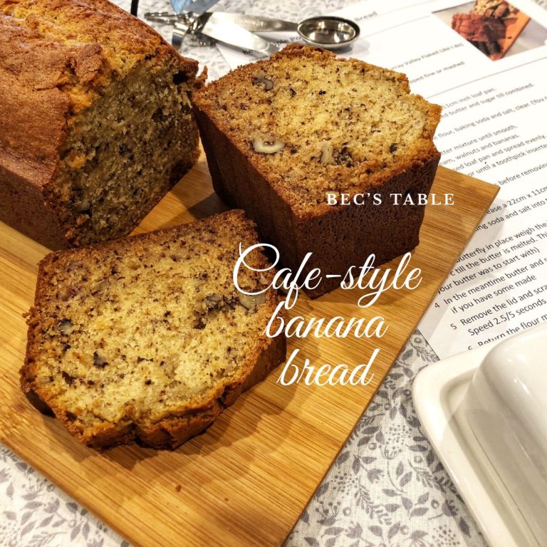 Banana bread sliced on a wooden board. with the recipe card in the back and a butter dish in the front