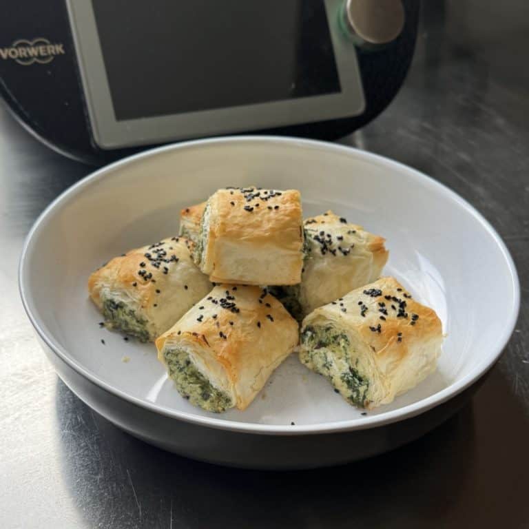 A white bowl with a stack of spinach, soft cheese and pine nut filo rolls sitting in front of a Thermomix